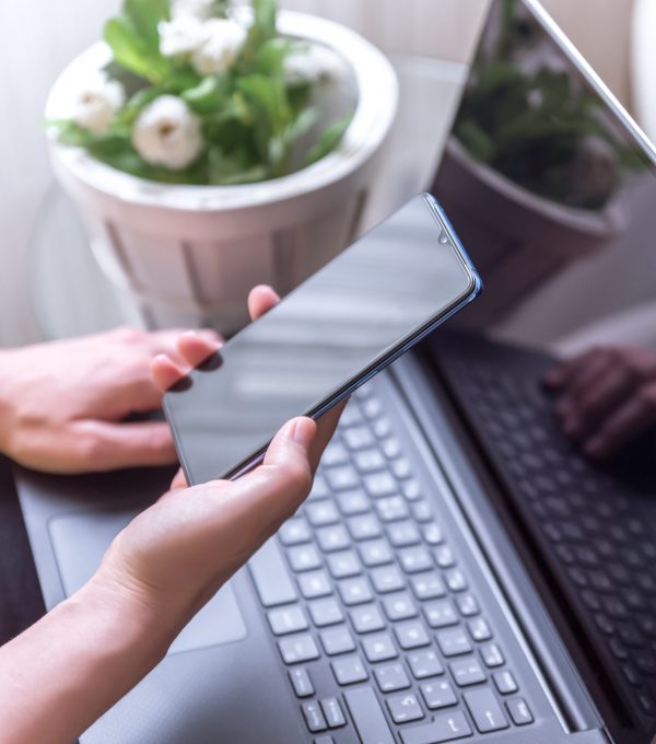 A vertical shot of a female hand holding a smartphone and laptop